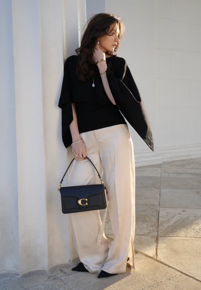 Mujer con top negro y pantalones anchos beige sosteniendo un bolso negro, de pie contra una pared blanca bajo una suave luz solar.