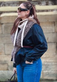 Veste en cuir noir avec col en fausse fourrure, écharpe en tricot crème, jeans en denim bleu et ceinture bordeaux, présentant une silhouette structurée.