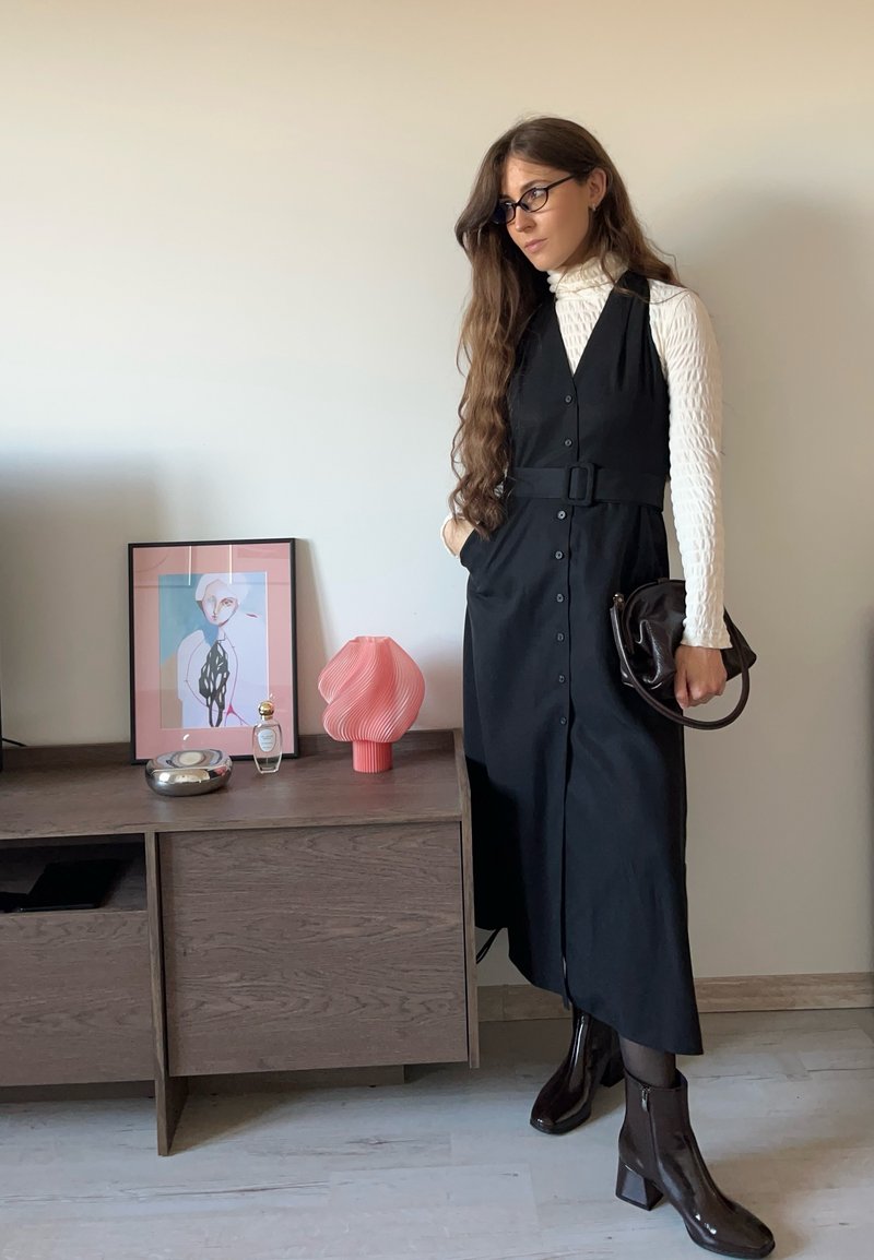 Vestito nero con bottoni e cintura, indossato sopra un dolcevita bianco con trama. Stivaletti scuri alla caviglia e una borsa marrone completano l'outfit.