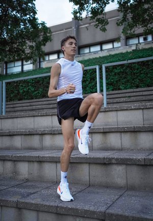 Top atlético sin mangas blanco, pantalones cortos negros y zapatillas de correr blancas con acentos en naranja. El sujeto está subiendo por unos escalones de piedra.