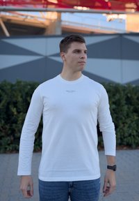 Λευκό πουκάμισο με μακριά μανίκια και στρογγυλή λαιμόκοψη. Διαθέτει ένα διακριτικό λογότυπο στο στήθος. Συνδυάζεται με μπλε τζιν, μπροστά από γεωμετρικό φόντο.