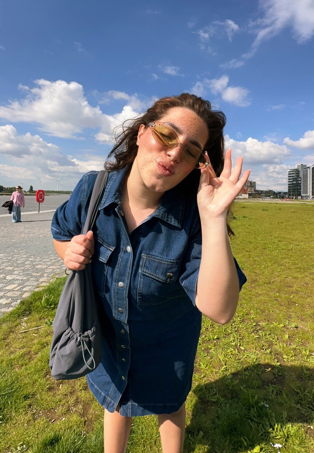 Robe chemise en denim à manches courtes, avec des boutons à l'avant et des poches. Cheveux ondulés, portant des lunettes de soleil à verres jaunes, tenant un sac à cordon gris.