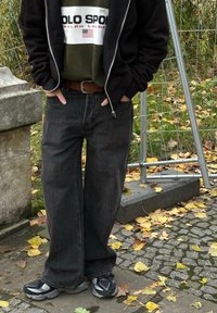 Czarna bluza z zamkiem na zamek założona na zieloną koszulę z logo, w połączeniu z luźnymi, ciemnymi dżinsami i szarymi trampkami. W tle widać jesienne liście i chodnik.