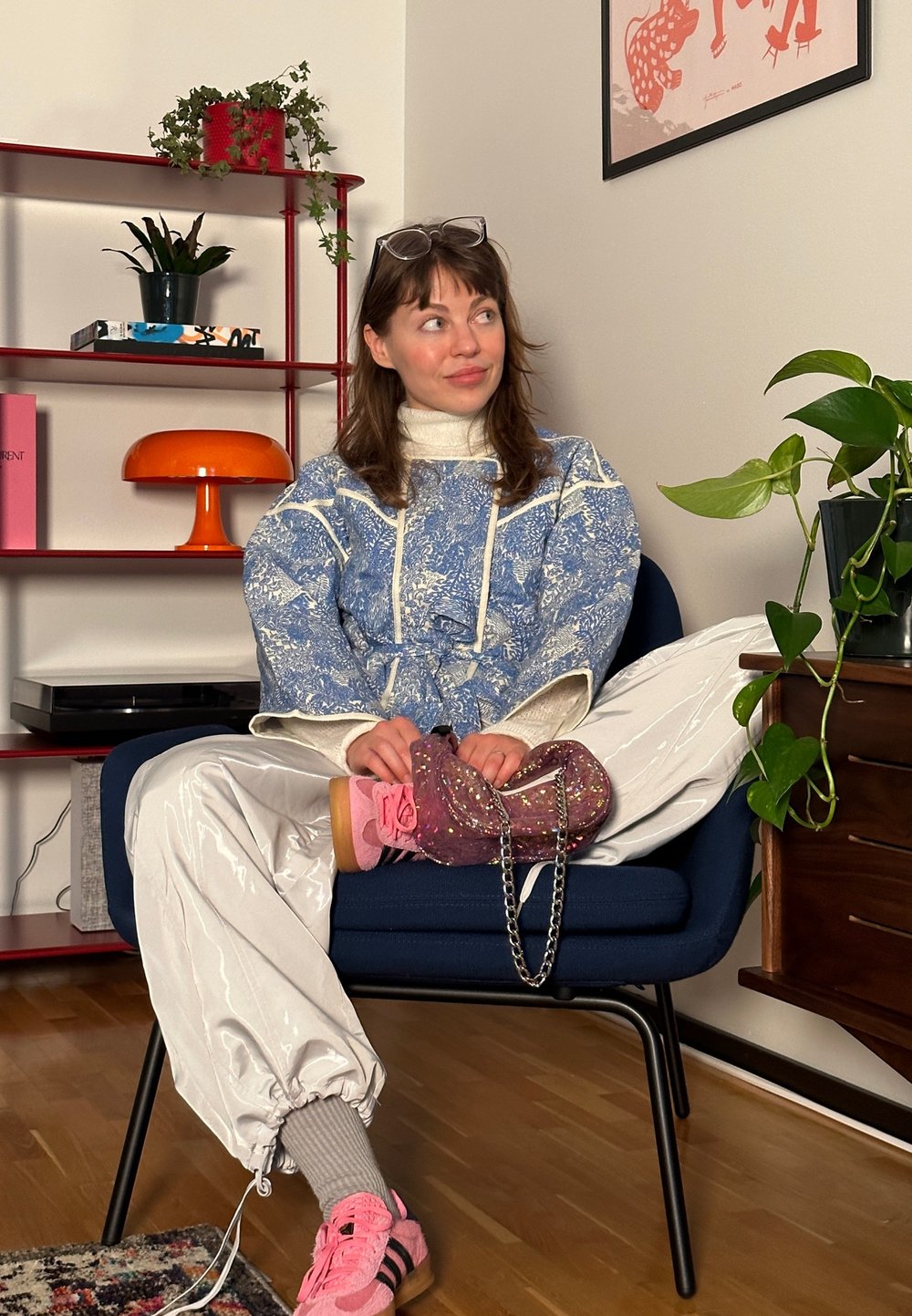 Μπλε πουκάμισο oversized με σχέδια και λευκές λεπτομέρειες, λαμπερό λευκό παντελόνι, ροζ αθλητικά παπούτσια και μια λαμπερή τσάντα. Εσωτερικός χώρος με φυτά και διακόσμηση.