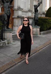 Svart ribbad klänning med franskant, kombinerad med svarta sandaler med remmar och en svart clutch. Accessoarer inkluderar mörka solglasögon.