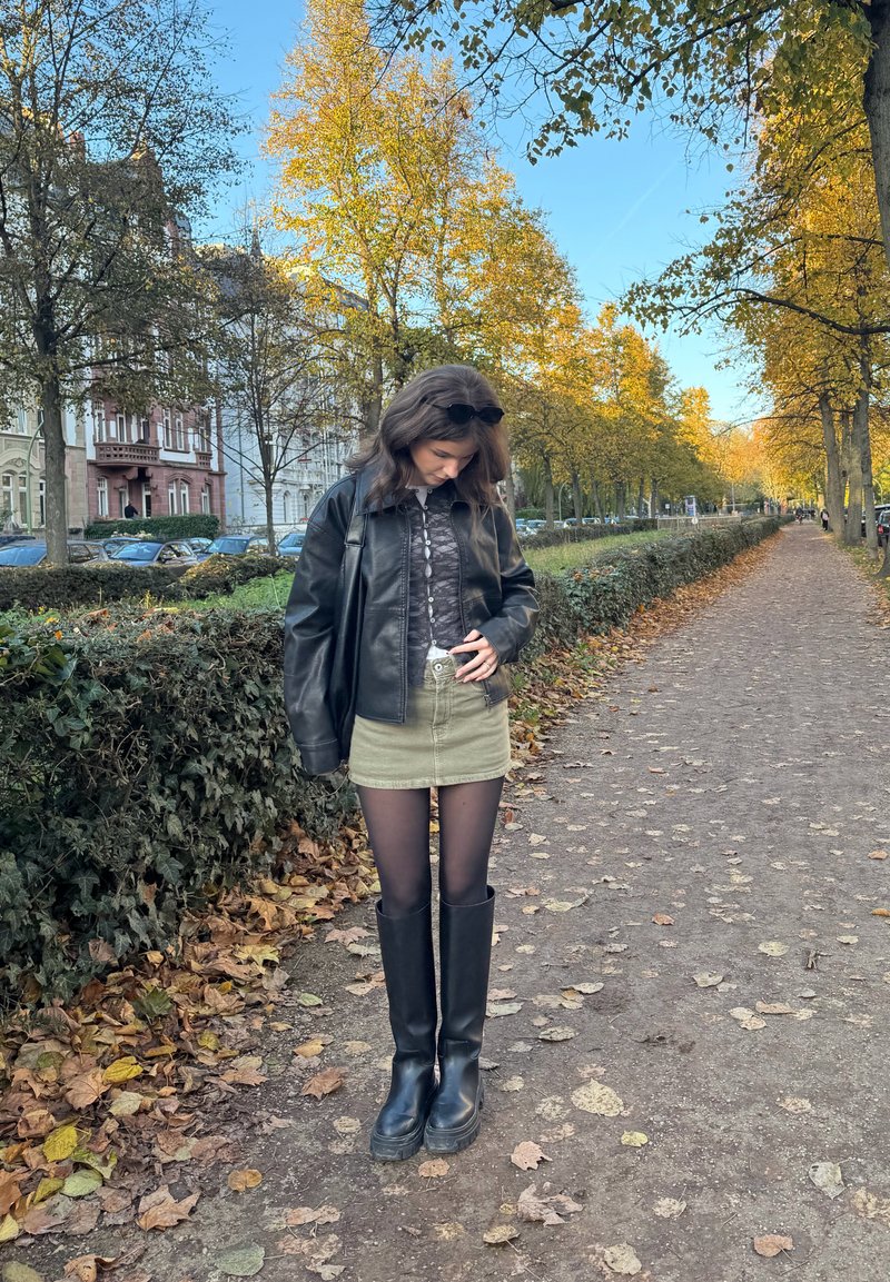 Veste en cuir noire, blouse blanche à motifs, jupe vert olive, collants noirs, bottes noires montant aux genoux, debout sur un chemin recouvert de feuilles avec des arbres.