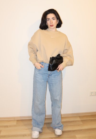 Mujer con cabello oscuro lleva sudadera beige, vaqueros azul claro, zapatillas blancas y sostiene un pequeño bolso negro frente a una pared lisa.