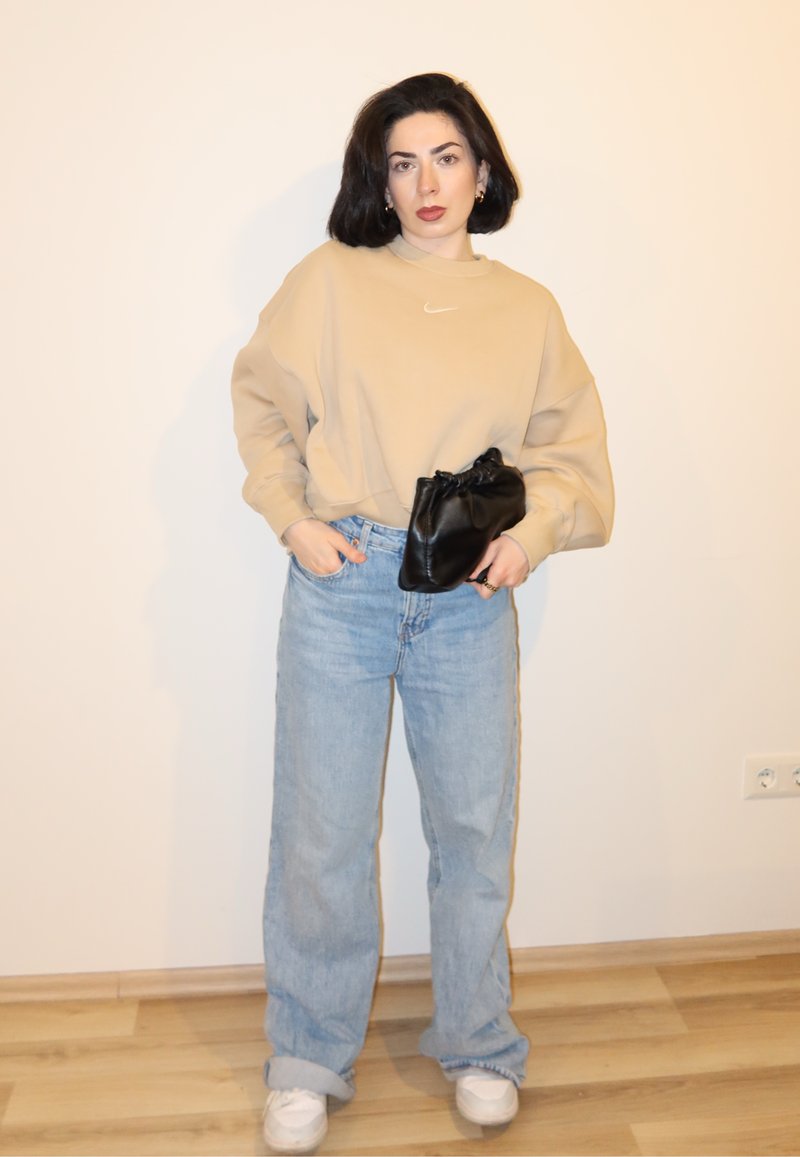 Γυναίκα με σκούρα μαλλιά φοράει μπεζ φούτερ, ανοιχτό μπλε τζιν, λευκά αθλητικά παπούτσια και κρατά μια μικρή μαύρη τσάντα μπροστά σε απλό τοίχο.