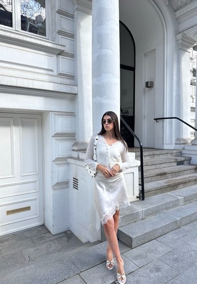 Mujer con cabello largo y oscuro, con cárdigan blanco, falda de satén con encaje, gafas de sol y bolso blanco, de pie junto a escalones y una columna de piedra.