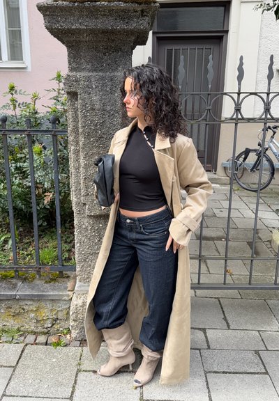 Mujer con cabello rizado que lleva un abrigo beige, top negro, jeans oscuros y botas beige, sosteniendo un bolso negro junto a un pilar de piedra y una cerca de metal.