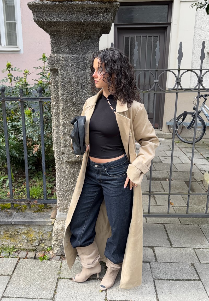 Kvinne med krøllete hår iført beige trenchcoat, svart topp, mørke jeans og beige støvler, holder en svart clutch ved en steinpilar og metallgjerde.