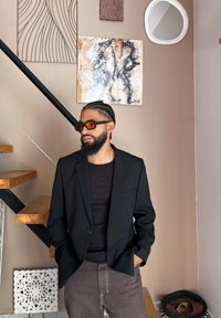 Μαύρο σακάκι πάνω από ένα σκούρο πουκάμισο, καφέ παντελόνι, γυαλιά ηλίου με πορτοκαλί απόχρωση. Ξύλινη σκάλα στο φόντο, διακοσμητική τέχνη τοίχου και καθρέφτης.