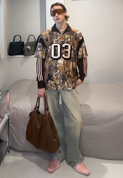Persona de pie con camiseta Adidas 03 camuflaje marrón sobre una camiseta de rayas de manga larga, vaqueros claros, zapatos rosas, sosteniendo un gran bolso marrón en interiores.