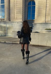 Veste en cuir noire avec accents en fourrure, mini-jupe noire et bottes noires brillantes montantes jusqu'aux genoux avec poignets en fourrure, sur fond d'un bâtiment en pierre.