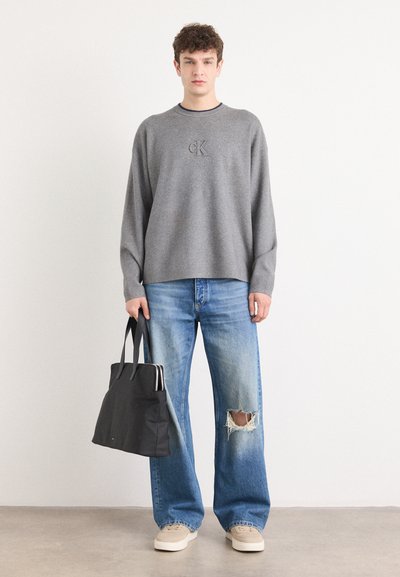 Suéter gris de punto con cuello redondo y logo, combinado con jeans de pierna ancha de color azul claro con una rodilla desgastada. Bolso tote negro.