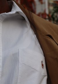 Biała koszula z bawełny z kieszenią, ozdobiona małym brązowym logo konia. Brązowa tkanina widoczna z kurtki nakładanej na koszulę.