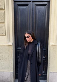 Long manteau noir, haut noir transparent et jupe à motifs. Sac noir mince avec bandoulière. Debout contre une porte sombre texturée.