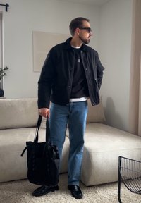 Homme portant une veste noire, un sweat-shirt noir, un jean bleu, des chaussures noires et des lunettes de soleil, tenant un sac fourre-tout noir dans un salon moderne.