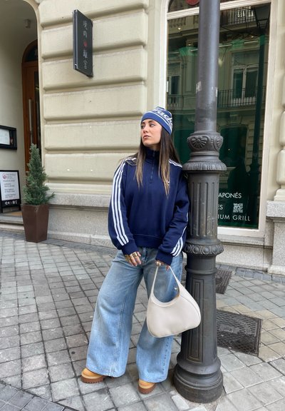 Sudadera navy con cremallera, rayas blancas y jeans de mezclilla sueltos. Bolso beige y zapatos de plataforma marrón. De pie junto a un farola.