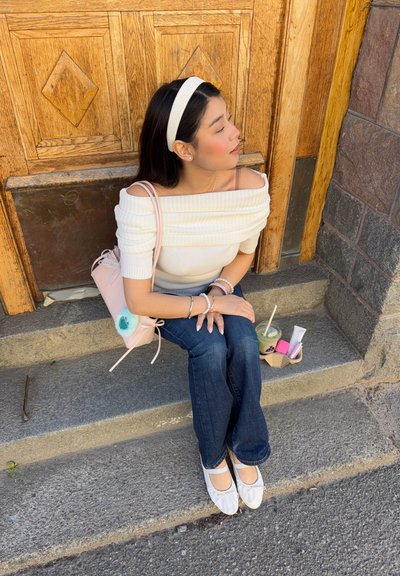 Joven mujer con top blanco de hombros descubiertos, vaqueros y diadema sentada en escalones de piedra junto a una bebida y artículos cosméticos.