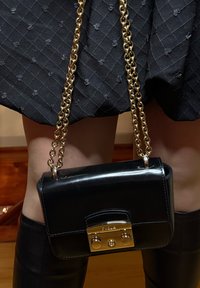 Sac à main en cuir noir avec une bandoulière chaîne dorée, forme rectangulaire, présentant un fermoir doré avec deux clous décoratifs et un logo sur le devant.