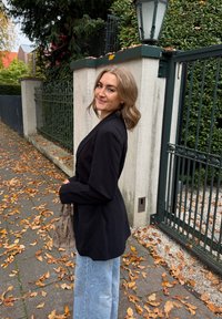 Sacou lung negru, blugi largi de culoare bleu deschis, ținând o geantă bej. Fundalul prezintă un perete de cărămidă și frunze de toamnă împrăștiate.