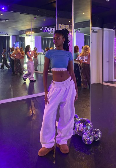 Una modelo con un top corto azul y pantalones de chándal blancos está de pie en el suelo de un estudio de baile. Hay bolas de discoteca reflectantes y espejos en el fondo.