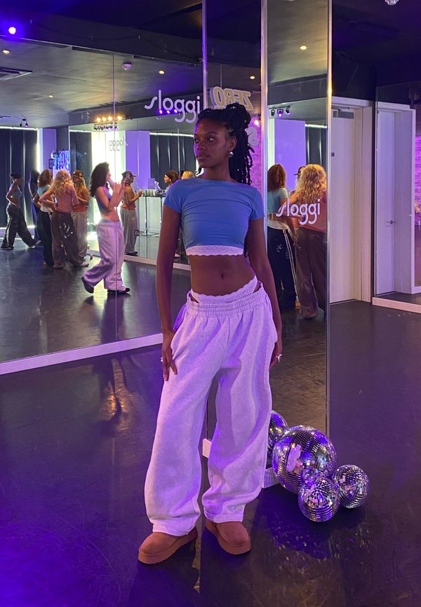Modella in un top corto blu e pantaloni tuta bianchi si trova su un pavimento di uno studio di danza. Palloni da discoteca riflettenti e specchi sono sullo sfondo.