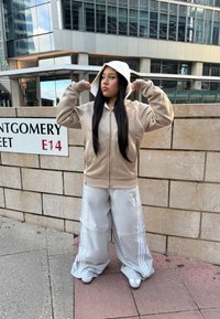 Felpa con zip di un beige chiaro con accenti bianchi, abbinata a pantaloni della tuta grigi oversize con strisce bianche. Sfondo di un muro di pietra.