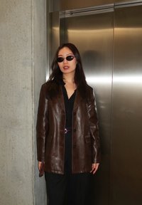 Femme portant des lunettes de soleil foncées et une veste en cuir marron, debout contre un mur en béton près d'une porte d'ascenseur métallique.