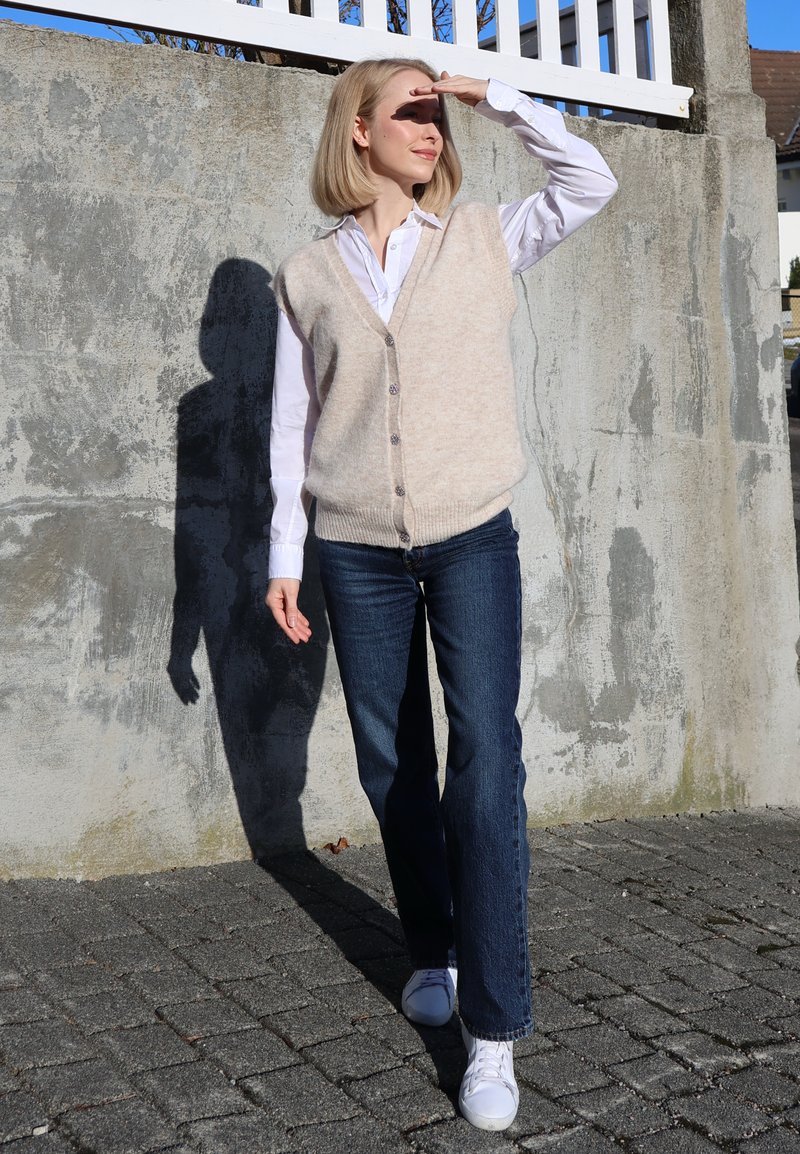 Femme en chemise blanche, gilet beige, jean bleu et baskets blanches, se protégeant les yeux avec la main en se tenant sur un sol pavé près d'un mur en béton.