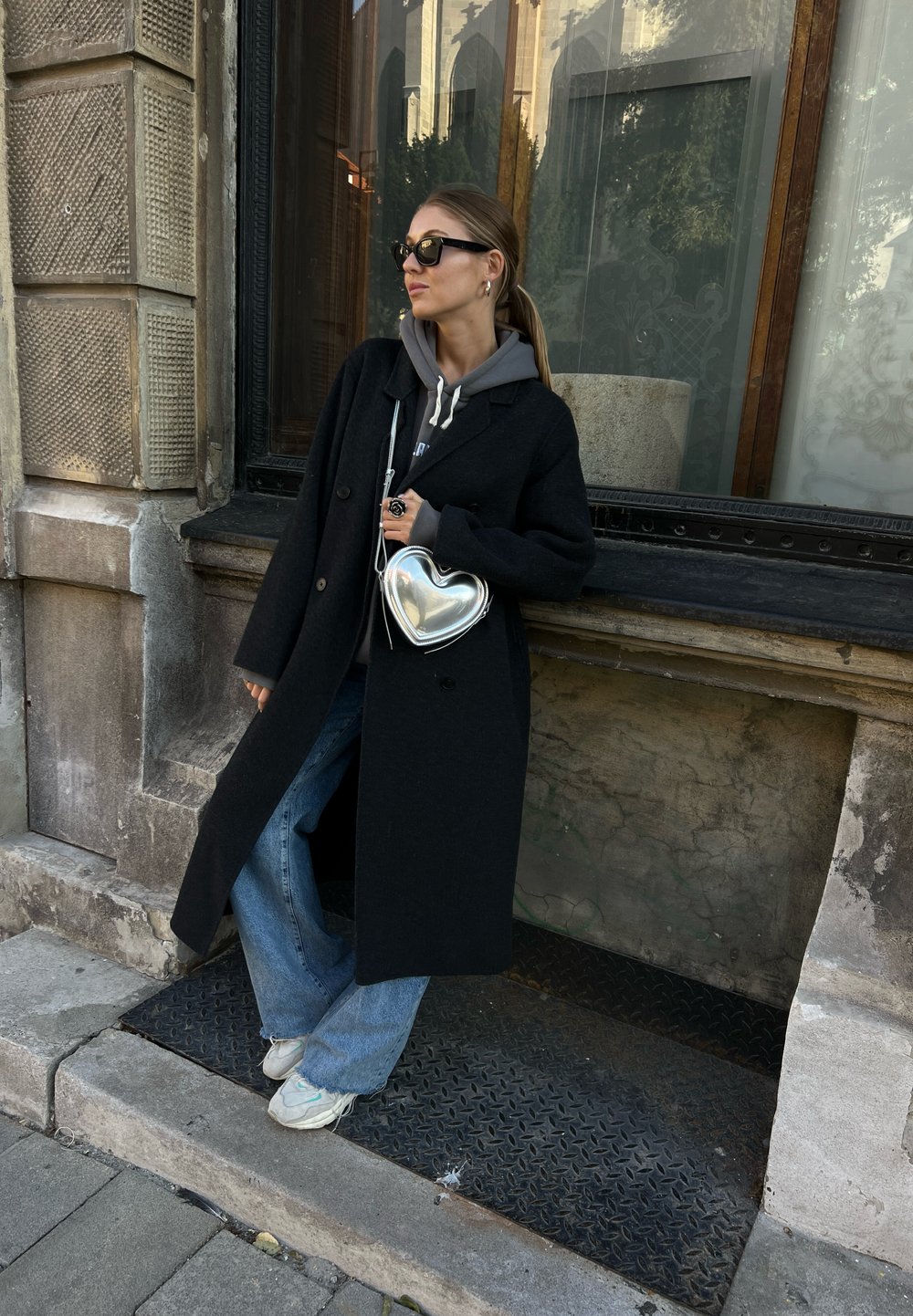 Lång svart kappa bärs över en grå hoodie, tillsammans med lösa blå jeans. En silverhjärtformad handväska och vita sneakers fulländar looken.