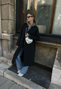 Lång svart kappa bärs över en grå hoodie, tillsammans med lösa blå jeans. En silverhjärtformad handväska och vita sneakers fulländar looken.