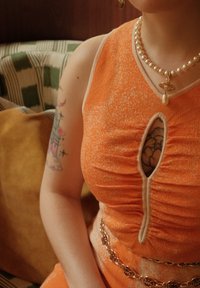 Een oranje gebreide jurk met een sleutelgat uitsnijding, geribbelde textuur en beige accenten. Een parelketting en een gouden ketting zijn zichtbaar.