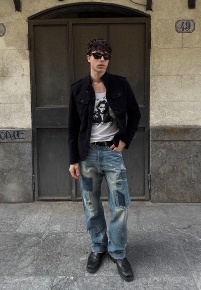 Chaqueta de mezclilla negra, camiseta gráfica blanca, jeans destrozados de mezclilla azul claro con parches, botas negras, de pie contra una pared gris texturizada.