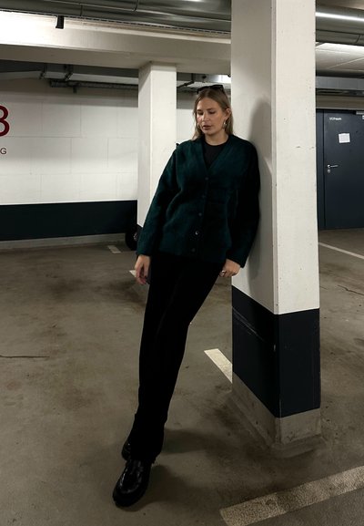 Cárdigan verde texturizado con botones, llevado sobre una blusa negra, combinado con pantalones negros. De pie contra un pilar blanco en un garaje.