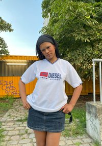 Bela kratka majica s kratkimi rokavi z modrimi detajli, ki vsebuje logotip "DIESEL DENIM DIVISION". Kombinirana s temno denim mini krilom.