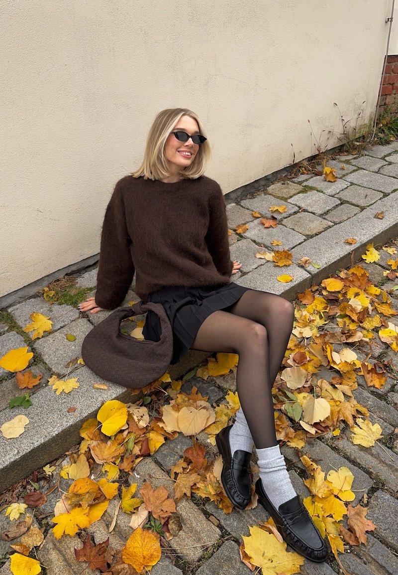 Hnědý, načechraný svetr, plisovaná černá sukně, silné černé punčocháče a mokasíny se světle šedými ponožkami. Hnědá pletená taška leží na zemi mezi podzimními listy.