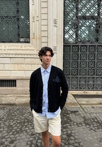 Cardigan noir avec boutons, présentant un design de planète ; chemise à rayures bleu clair, boutonnée en dessous ; shorts crème. Cadre extérieur en pavés.