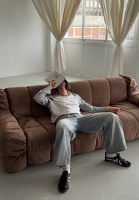 Muž opírající se o hnědou pohovku s knihou zakrývající tvář v světlé místnosti s velkým oknem a krémovými závěsy.