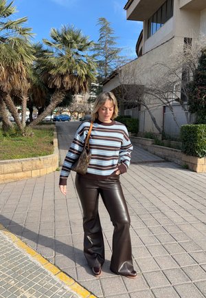 Mujer con suéter a rayas, pantalones de cuero marrón oscuro y bolso marrón, caminando por una acera pavimentada soleada con palmeras cerca.