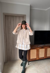 Γυναίκα που διορθώνει μαύρο καπέλο, φορώντας μπεζ πουκάμισο με καρό σχέδιο, εκρού γιλέκο από φλις, μαύρο κολάν και πράσινες αδιάβροχες μπότες, στέκεται μέσα σε εσωτερικό χώρο κοντά σε επιπλο τηλεόρασης.