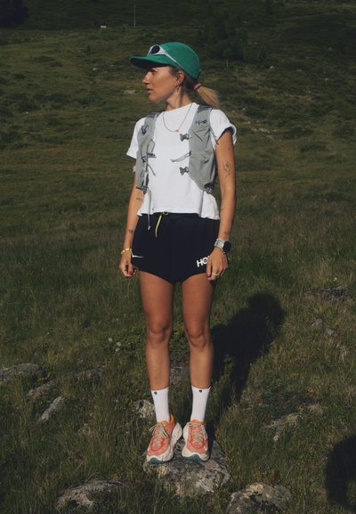 Conjunto atlético que incluye un chaleco gris, una camiseta blanca corta, pantalones cortos negros con logo, calcetines blancos y zapatillas de correr coloridas. Gorra verde y gafas de sol.