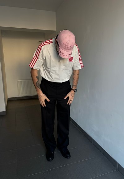 Camisa blanca con rayas rojas y logo, pantalones anchos negros, gorra rosa con texto y joyería plateada. Entorno interior con suelo de baldosas.