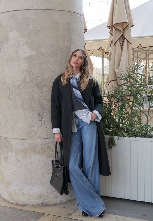 Ung kvinne kledd i svart frakk, stripete skjorte, blått slips og vide jeans, holder en svart håndveske og lener seg mot en stor steinsøyle utendørs.