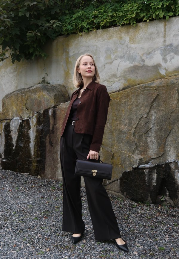 Mujer con chaqueta marrón y pantalones negros sostiene un bolso negro, parada sobre grava frente a un muro de piedra con vegetación encima.