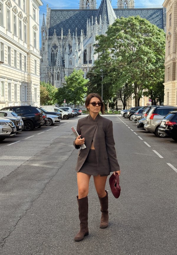 Mujer con gafas de sol que lleva una blazer marrón, falda y botas caminando por una calle de la ciudad con una catedral histórica y coches aparcados al fondo.