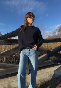 Γυναίκα που φοράει γυαλιά ηλίου, μαύρο πουλόβερ και μπλε τζιν στέκεται έξω δίπλα σε μια ξύλινη περίφραξη κάτω από καθαρό γαλάζιο ουρανό.