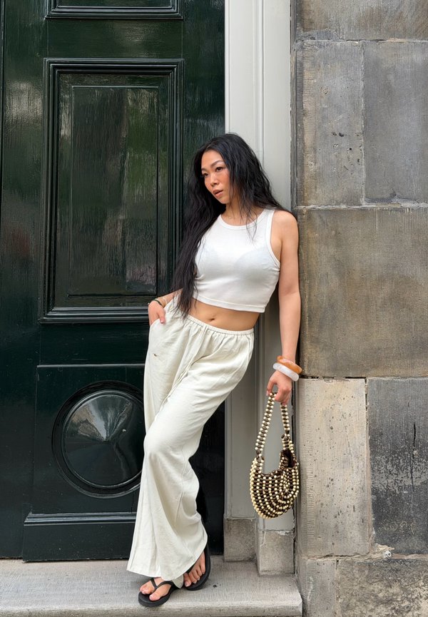 Donna con crop top bianco e pantaloni si appoggia al muro di un edificio tenendo una borsa di perline, in piedi vicino a una grande porta verde scuro.