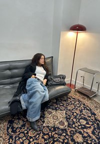 Črno usnjena zofe, modre prevelike kavbojke in črni gležnjarji. Sive ročne torbice počivajo na naročju. Vzorec preproge in moderna svetilka dodajata detajle.